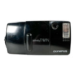 An Olympus Infinity Twin black film camera with a visible flash unit attached, photographed against a light gray background.
