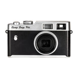 A retro-style compact camera with a black textured body and silver metal accents, featuring the text 'Camp Snap PRO' on the top left. The camera is photographed on a clean, light beige background with a soft reflection below.