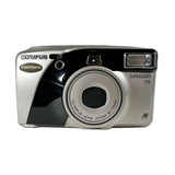 An Olympus SuperZOOM 115 QuartzDate instant camera with a silver and black body, shown from a front three-quarter angle on a light gray background.
