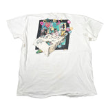 A white short-sleeve graphic t-shirt laid flat on a light gray surface. The front of the shirt features a large, colorful square graphic print depicting a surreal, cluttered scene with a bed, balloons, and the word 'DONT' at the top.