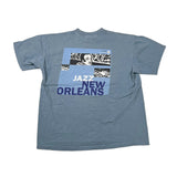 A faded blue short-sleeve t-shirt laid flat on a light beige background, featuring a graphic print on the back with the words 'JAZZ NEW ORLEANS' and a black-and-white portrait collage.