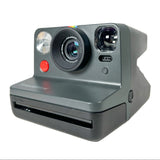 A Polaroid Instant Mini camera in dark gray, shown from a three-quarter angle on a light gray background. The camera features a prominent lens, a red shutter button, and a built-in flash.
