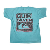 A vintage turquoise short-sleeve t-shirt laid flat on a white background, featuring a large distressed black and pink graphic on the back with the text 'QUIK SILVER' and a wave logo.