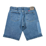 A pair of blue denim shorts shown from the back, laid flat on a white fabric background. The shorts feature classic five-pocket styling with a distinctive red tab on the back waistband and a large patch pocket on the right leg.