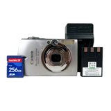 A silver Canon PowerShot S2500 compact camera shown with a blue SanDisk 256MB SD card on the left, a black Canon battery charger on the right, and a black Canon rechargeable battery in front of the charger, all photographed on a light gray background.