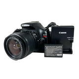 Canon EOS Rebel T3 DSLR camera with a zoom lens, shown with a Canon battery charger and a replacement battery on a light gray background.