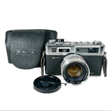 A vintage Yashica camera with a silver and black body, shown alongside its black textured leather case and lens cap on a white background.