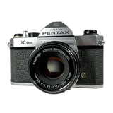 A vintage Pentax K1000 film camera with a black textured body, silver metal dials, and a black lens, photographed on a light gray background.