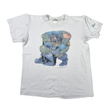 A white short-sleeve graphic t-shirt laid flat on a light gray background. The front features a large, distressed illustration of Batman crouching in a chair, colored in shades of blue, green, and yellow.
