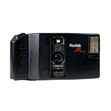 A vintage black Kodak S- series single-lens reflex camera with a front view, featuring the Kodak logo and a flash unit on the side, set against a plain light gray background.
