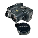 An Olympus SW2000 SWAP camera in black, shown with its matching black lens cap on a white textured background.