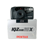 A black Pentax IQZOOM 60X compact camera placed on top of its original gray packaging box, photographed against a neutral light gray background.