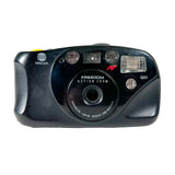 A black Minolta Freedom Action Zoom QD compact camera photographed from the front against a light gray background.