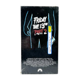 A sealed VHS case for the movie 'Friday the 13th Part 2' standing upright against a light gray studio background. The cover is predominantly black with bright blue line-art figures, white title text, and red 'PART 2' text, plus a yellow-and-white barcode sticker on the right side.