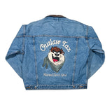 A blue denim jacket shown from the back, featuring a large embroidered Tasmanian Devil graphic with the text 'Outlaw Tax' and 'Warned Since 1964' on a white background.