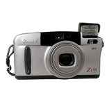 A Canon Z135 compact digital camera in silver and black, featuring a built-in flash and wrist strap, photographed on a light gray background.