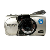 An Olympus OStylus 200M 115 zoom camera in silver with a black lens, featuring a blue 'Luminance' sticker and a black 'QuartzDate' sticker on the front, set against a neutral gray background.
