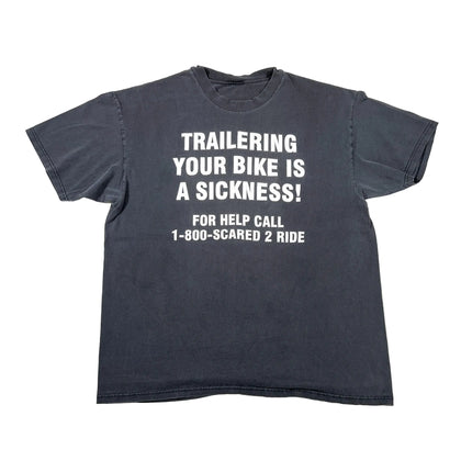 A faded black short-sleeve t-shirt with bold white text that reads 'TRAILERING YOUR BIKE IS A SICKNESS! FOR HELP CALL 1-800-SCARED 2 RIDE', laid flat on a white background.