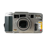 An Olympus Accura Verios compact camera in silver and black, featuring a visible yellow 'Mature Date' sticker on the front, photographed on a light gray textured background.