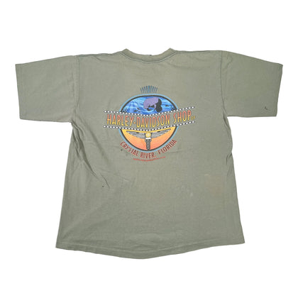 An olive green short-sleeve t-shirt laid flat on a white surface, featuring a large circular graphic on the back with the text 'Harley-Davidson Shop' and 'Crescent River, Florida'.