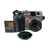 An Olympus Cmedia CCD camera in silver and black, shown with its black lens cap and a 16MB SD card on a light gray background.