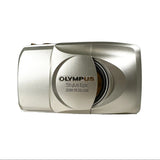 An Olympus Stylus Epic Zoom 170 Deluxe digital camera in metallic silver, photographed on a light gray textured surface with a neutral gray background.