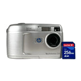 A silver HP Photosmart 320 compact digital camera shown next to a blue SanDisk 256MB SD card on a light gray background.