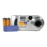 A silver Sony Cyber-shot digital camera shown with two orange AA batteries and a blue Sony memory stick on a light gray background.