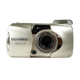An Olympus Stylus 120 compact digital camera in metallic silver, photographed from the front against a light gray background.