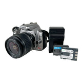 A Canon EOS Rebel xt SLR camera with a zoom lens, shown alongside a black battery charger and two Canon NP-F batteries on a light gray background.