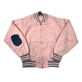A light pink vintage bomber jacket with gray and white striped ribbed collar, cuffs, and hem, featuring a dark blue patch on the right sleeve. The jacket is laid flat on a white background.