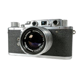 A vintage Nippon Kodo Nikkor 50mm f/2.8 camera lens attached to a silver and black rangefinder camera, photographed on a clean white textured surface.