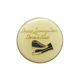 Round gold pin with shoe design and 'Bruce Springsteen Born to Run' text on a white background