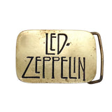 A vintage Led Zeppelin belt buckle in a warm gold-toned metal, featuring the band's name in black engraved lettering and a matching metal belt loop, shown on a textured white background.