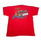 A red short-sleeve graphic t-shirt laid flat on a white background. The front of the shirt features a colorful retro-style graphic with the text 'BAD DOG' above a cartoon palm tree, surfboards, a beach chair, and a sign.