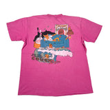 A bright pink short-sleeve graphic t-shirt laid flat on a white background. The front features a colorful cartoon illustration of a man sitting on a blue couch with a cat on his lap, accompanied by the text 'BAD DOG PROLOCONDO COUCH' and 'LOVE' in stylized lettering.