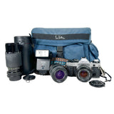 A blue fabric camera bag with a black strap and white script logo, displayed with vintage camera gear including a silver and black Canon SLR camera, a Vector flash, a lens, and a black cylindrical case on a white studio background.
