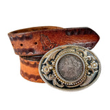 A brown tooled leather belt with a large oval silver-tone buckle featuring an embossed Morgan silver dollar coin in the center, displayed on a white background.