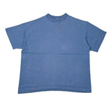 A faded medium blue short-sleeve crewneck t-shirt laid flat on a white surface, with the bottom hem slightly wrinkled and black pants visible at the bottom edge.