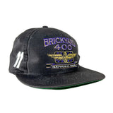 A black snapback cap featuring embroidered 'BRICKYARD 400' in purple and gold, with a winged '400' logo and 'INAUGURAL RACE' text on the front, and white embroidered numbers on the side.