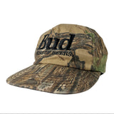 A Bud King of Beers baseball cap featuring a woodland camouflage pattern in shades of brown, green, and beige, with black embroidered lettering on the front.