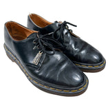A pair of black leather lace-up oxford shoes with yellow contrast stitching and a visible 'DR. MARTIN' tag, photographed on a white fabric background.