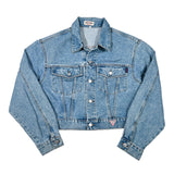 A light blue denim trucker jacket laid flat on a white background, featuring a button-up front, chest flap pockets, and a small pink-and-white Guess logo patch near the lower left hem.