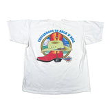 A white short-sleeve graphic T-shirt laid flat on a light gray surface. The back of the shirt features a colorful western-themed print with the text 'CROSSROADS OF ROCK 'N ROLL' in blue around an illustration of a cowboy hat and red boots.