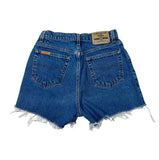 Back view of a pair of blue denim shorts with a raw, frayed hem, featuring a tan leather patch on the back waistband and a small orange tag on the right pocket.