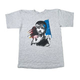 A heather gray short-sleeve t-shirt laid flat on a white surface, featuring a graphic print of a girl with black hair, a pink heart, and a blue paint-stroke background.