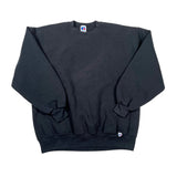 A black crewneck sweatshirt laid flat on a white surface, showing the front chest, ribbed cuffs, and a small brand tag near the lower hem.