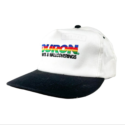A white baseball cap with a black visor, featuring a colorful embroidered logo on the front that reads 'DURON WITS & WALLCOVERINGS'.