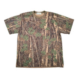 A short-sleeve crewneck t-shirt featuring a forest camouflage pattern with brown tree trunks, beige branches, and green leaves. The shirt is laid flat on a white surface.