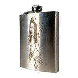 A stainless steel hip flask with a engraved illustration of a woman and a small skull detail, shown on a neutral light gray background.